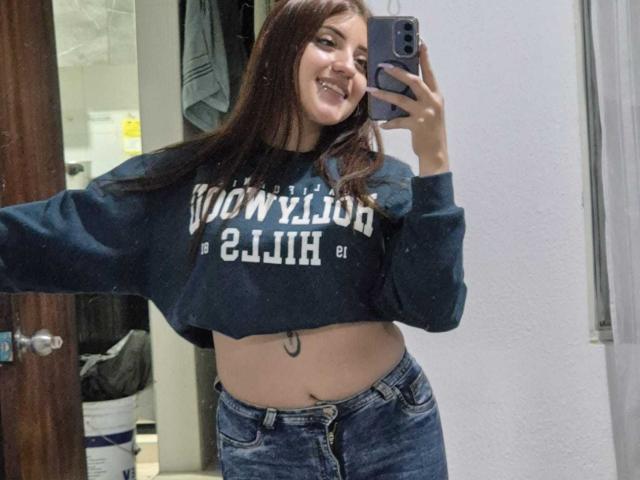 MaylinSanchez with long brown hair, smiling, wearing a dark blue cropped sweater, showing her belly button tattoo, and blue jeans, taking a mirror selfie.