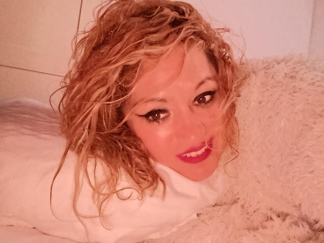 NatashaChaus with curly blonde hair, pink lipstick, and white eyeliner, lying on a fluffy white pillow, smiling at the camera, wearing a white shirt, looking sexy.