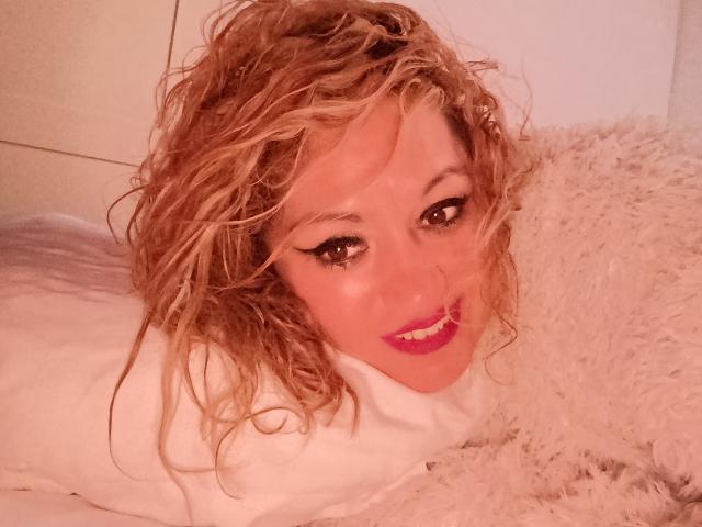 NatashaChaus with curly blonde hair, pink lipstick, and white makeup, lying on a fluffy white pillow, smiling at the camera, looking sexy.