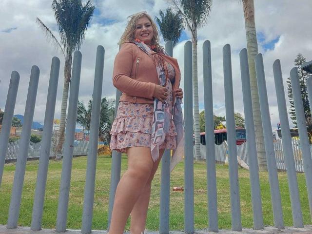 NatashaChaus with blonde hair, pink dress, floral scarf, and brown jacket, standing in front of a tall, gray, metal fence, smiling, with palm trees and a white fence behind.