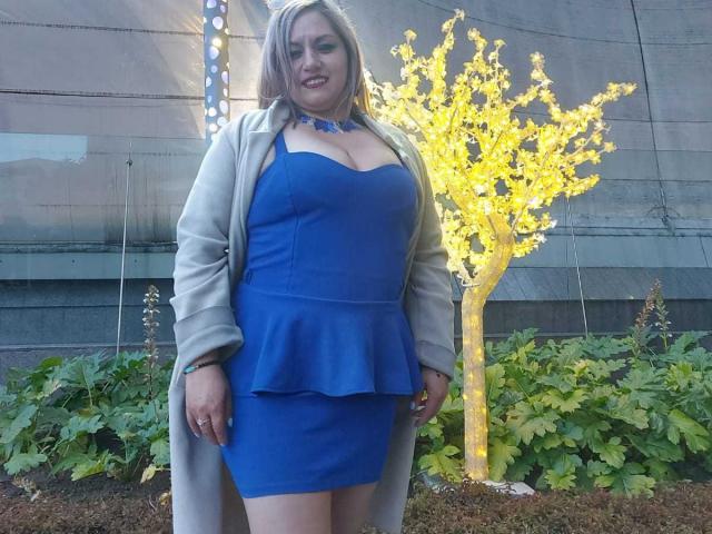 NatashaChaus with big tits in a blue dress, flirty ruffle, gray coat, blonde hair, smiling, standing in front of a yellow illuminated tree, green plants below.