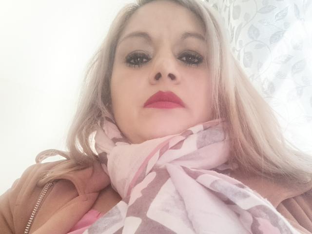 NatashaChaus with pink lipstick, blue eyes, and long blonde hair, wearing a brown jacket and pink-and-white scarf, has her nose and lips prominently in the foreground.