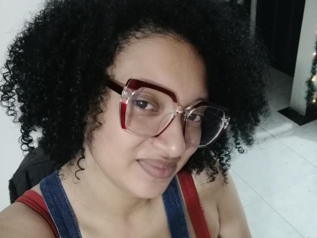 VioletaSwitch has curly black hair, red-framed glasses, and is wearing a red and blue tank top, showing a subtle smile with her tits slightly visible.