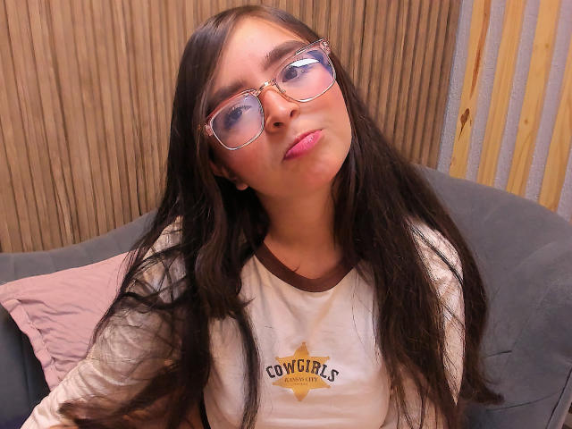 EvaMcAllister with long brown hair, wearing glasses, pink lipstick, and a white "Cow City" shirt, looking slightly up with a relaxed expression. Boobs not visible, pussy not visible.