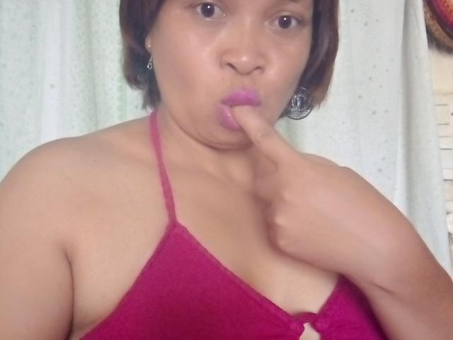 Rahel69, com pele morena, cabelo castanho curto e batom roxo, chupando o dedo indicador, vestindo uma blusa vermelha de alça, mostrando seus pequenos seios e ombros nus..