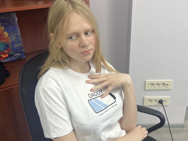Laliina with blonde hair, white shirt, and "Daddy" graphic, sitting in chair, hand on chest, looking to the side, slight red marks on cheek, electrical outlet behind.