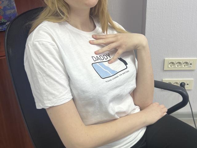 Laliina with blonde hair, wearing a white t-shirt, sitting on a black chair, hand touching her chest, showing her tits, in a room with a gray wall.