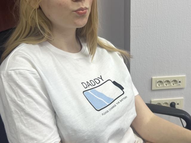 Laliina with blonde hair, pouty lips, wearing a white t-shirt with a battery graphic and text, sitting in a chair, showing her tits slightly, in a room.