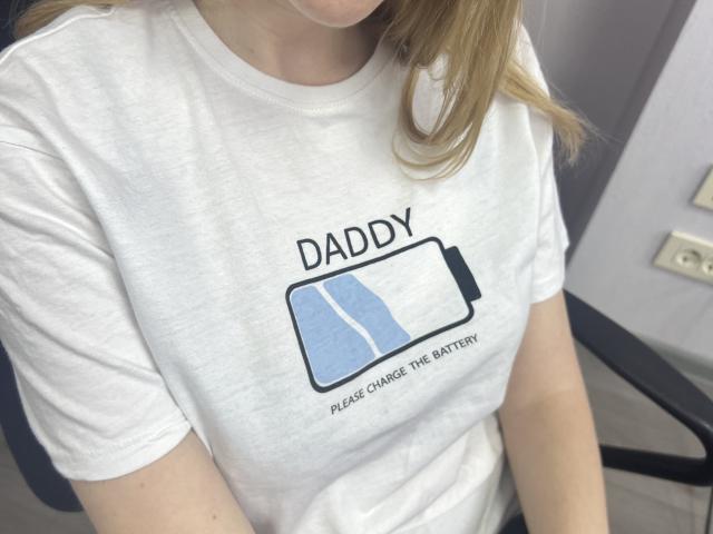 Laliina's white t-shirt with a battery graphic and text, showing her upper chest and arms. She has blonde hair, and the shirt's texture is slightly rough.