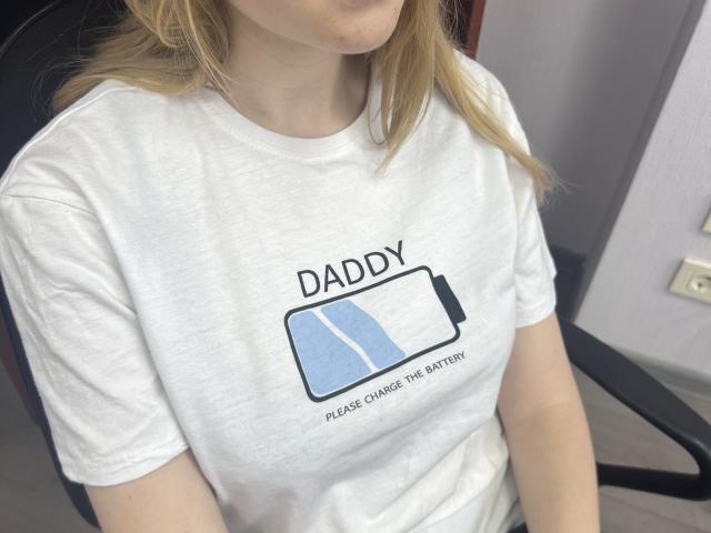 Laliina with blonde hair, wearing a white t-shirt showing a battery with text, sitting on a chair, her small tits slightly visible under the fabric.
