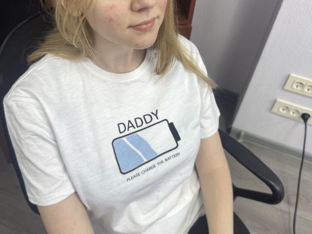 Laliina with blonde hair, wearing a white t-shirt, sitting in a chair, has small, pale tits, light skin, subtle red marks on her face, and a slightly shy smile.