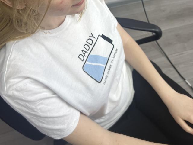 Laliina with blonde hair, wearing a white t-shirt, sitting in a chair. Her shirt has a blue battery graphic with "DADDY" above it. Her ass is partially visible.