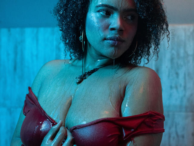 AliceJelanni met nat, krullend haar, huid met een blauwachtige tint, en een rood, doorweekt topje, die haar grote, glanzende borsten vasthoudt, met waterdruppels, met een serieuze blik, en die oorringen en een ketting draagt..