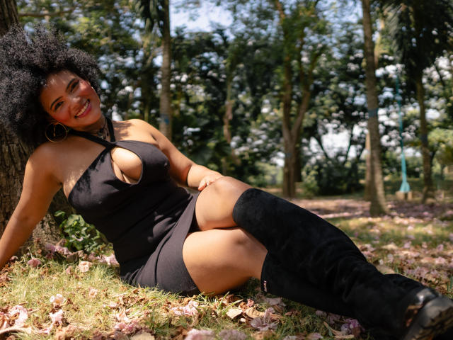 Alice Jelanni, con cabello negro rizado, lápiz labial rojo y aros grandes, sentada en el césped con un vestido negro con un corte que muestra sus senos, y botas negras hasta la rodilla..