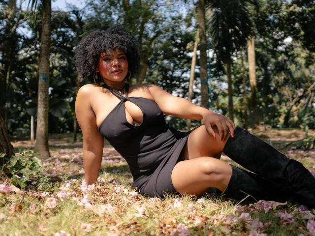 AliceJelanni with curly black hair, wearing a black halter dress with a cutout, and black knee boots, sitting on grass with pink petals, smiling at the camera.