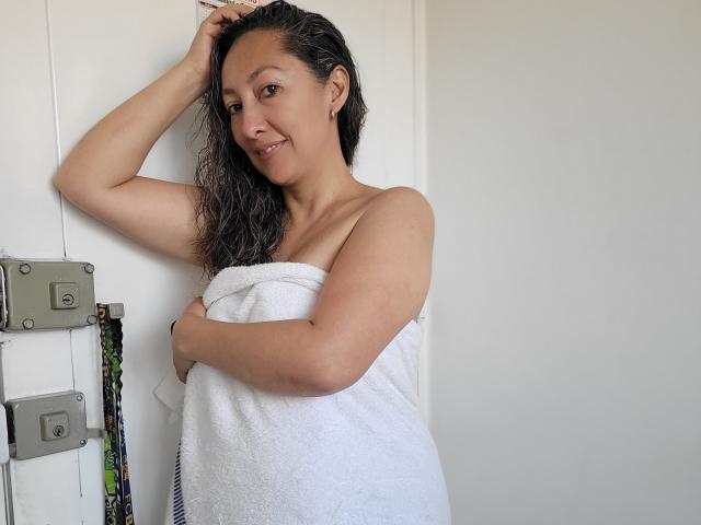 Sandhy: Capelli scuri e umidi, sorridente, con un asciugamano bianco che copre i seni e i glutei scoperti, in piedi di fronte a una porta bianca, con un braccio alzato, che appare carina e rilassata..