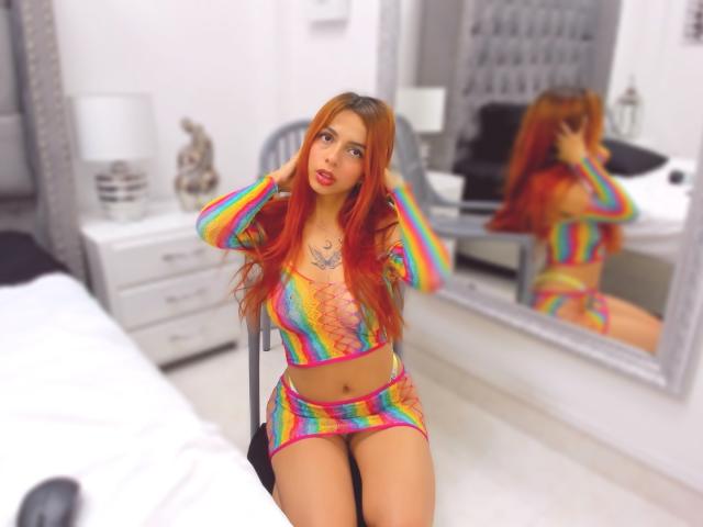 SamantaCarter with long red hair, wearing a rainbow-striped crop top and mini skirt, sitting in a bedroom, hands in her hair, reflecting in a mirror, showing her tits.