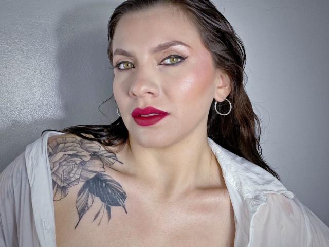 KarlaMorriss with wet brown hair, red lipstick, green eyes, hoop earrings, white shirt, and a tattoo of roses on her left shoulder. Her tits are visible.