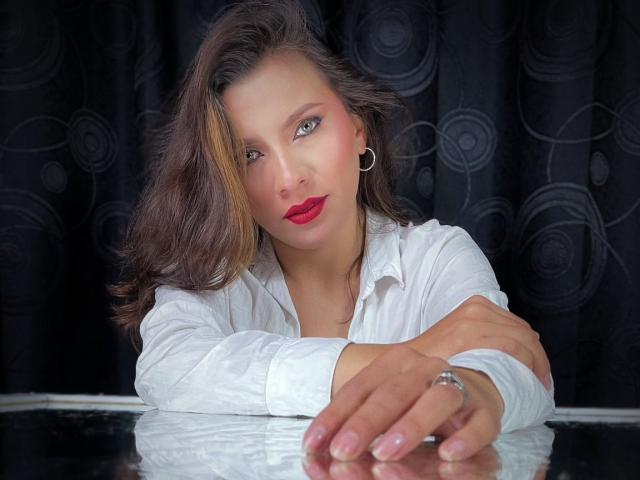 KarlaMorriss has wavy brown hair, red lipstick, black ear gauges, and wears a white shirt, leaning on a shiny surface with her hand, showing a ring.