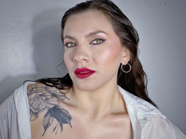 KarlaMorriss with wet brown hair, red lipstick, green eyes, white shirt, hoop earrings, and a tattoo of roses on her right shoulder. Her tits are slightly visible.