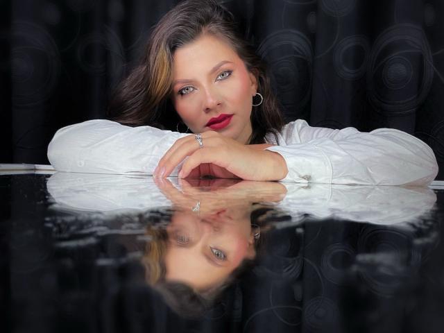 KarlaMorriss with red lipstick, white shirt, and ring, leaning on a reflective black surface, showing her reflection, with dark hair and hoop earrings, looking at the camera.