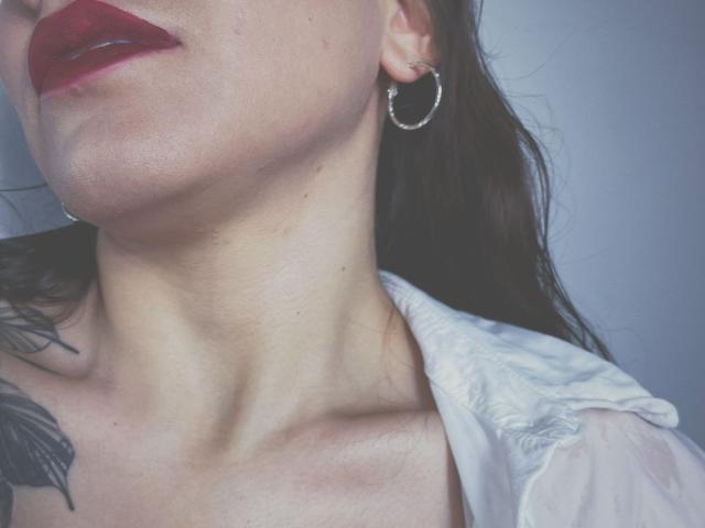 KarlaMorriss's neck and upper chest with red lipstick, hoop earring, white shirt, and black feather tattoo. Her skin is light, and she has a slight tan.