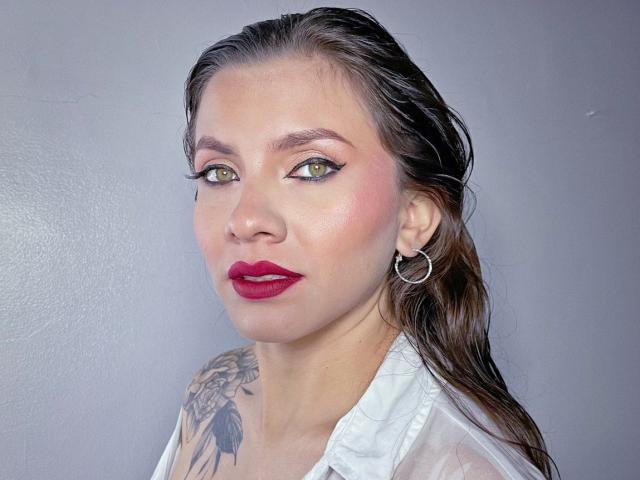 KarlaMorriss has wet brown hair, green eyes, red lipstick, white shirt, hoop earrings, and a tattoo on her right shoulder. Her tits are partially visible.