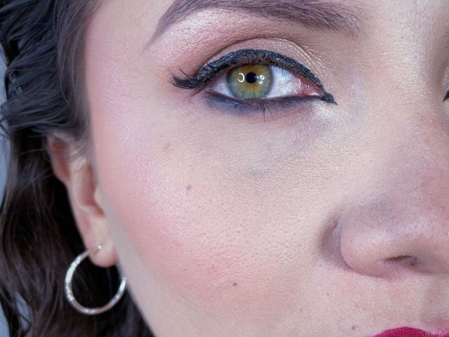 KarlaMorriss's eye is yellow with black eyeliner, thick lashes, and a slight brow. She's wearing a silver hoop earring and has a pinkish skin tone with small moles.