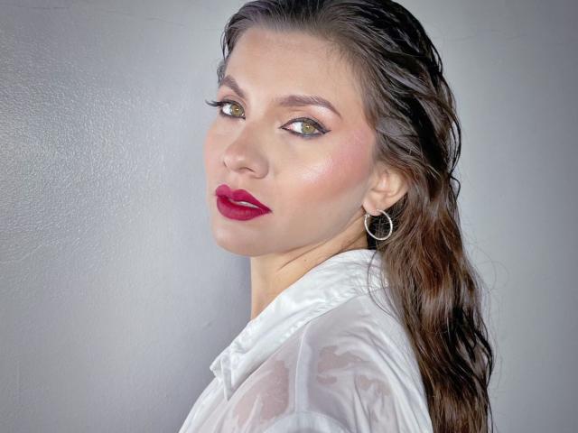 KarlaMorriss with wet brown hair, green eyes, red lipstick, white sheer top, hoop earrings, looking over shoulder, slightly smiling, soft light on face.
