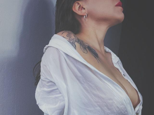 KarlaMorriss with wet black hair, red lips, white shirt open to reveal cleavage, and a floral tattoo on her right shoulder. Her tits are partially visible.