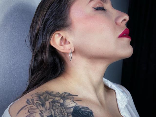 KarlaMorriss with wet brown hair, pink lips, and white shirt, showing off a floral tattoo on her shoulder, wearing silver earrings, looking up with eyeliner.