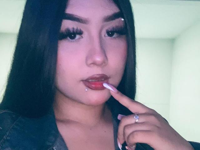 DavidAndLaura with long black hair, wearing red lipstick, touching her lips with a finger, showing a nose piercing, and wearing a denim shirt, with a ring on her finger.