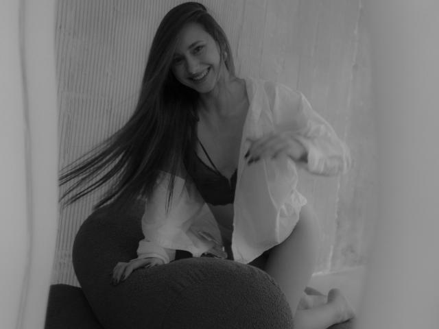 Black-and-white photo of LaraSweetyHot with long hair, smiling, wearing an open shirt, dark bra, and no pants, kneeling on a textured couch, showing her tits and ass.