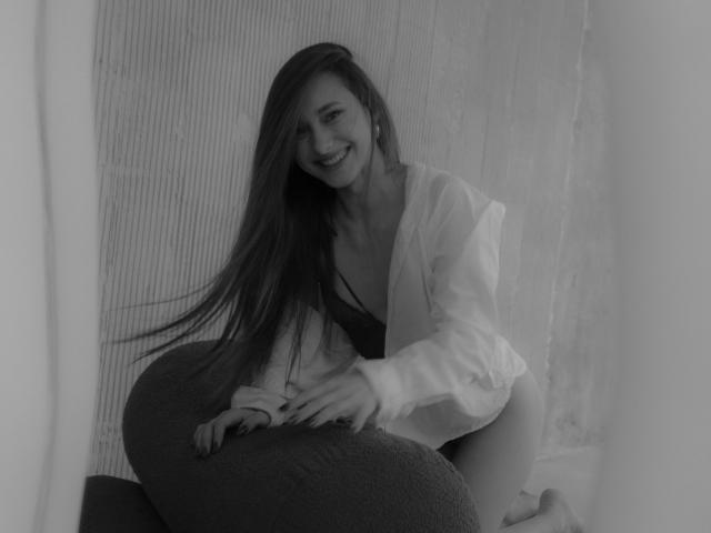 Black and white photo of LaraSweetyHot with long hair, smiling, wearing an open shirt, kneeling on a pillow, showing her tits and ass, looking cute and sexy.