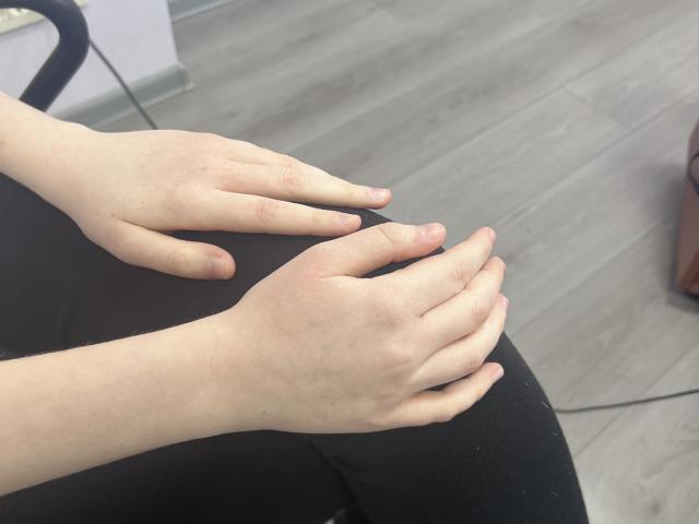Laliina's pale hands resting on her black pants, fingers slightly spread, showing light skin and minimal texture. She's sitting on a wooden floor with a chair visible.