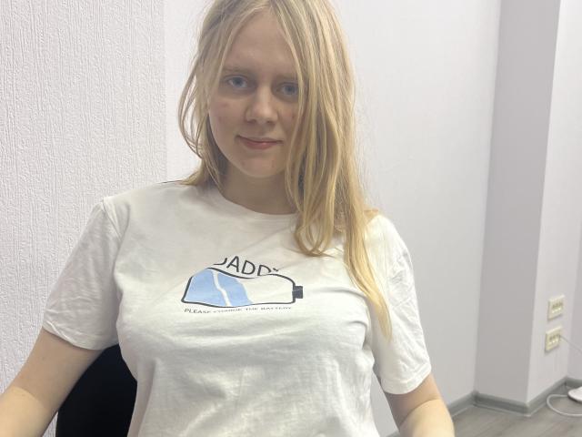 Laliina with blonde hair, wearing a white t-shirt, has small tits and a subtle smile, standing in a room with light purple walls. She's slightly chubby and cute.