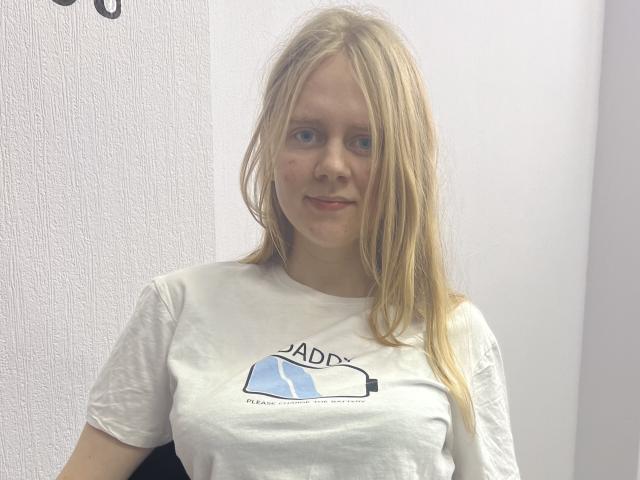 Laliina with blonde hair, blue eyes, slight smile, wearing white t-shirt, small tits, fair skin, standing against white wall, looking at camera, casual, relaxed, slight redness on cheek.