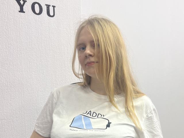Laliina has long blonde hair, a white t-shirt with a blue and black graphic, and a slight smile. Her tits are small, and she's standing against a white wall.