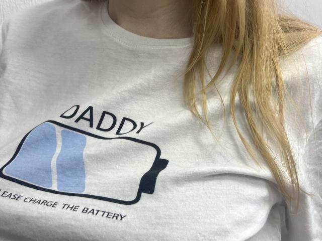 Laliina's white t-shirt with a blue and white battery graphic, labeled "Daddy" in black text, showing her small, perky boobs. Long blonde hair partially visible.
