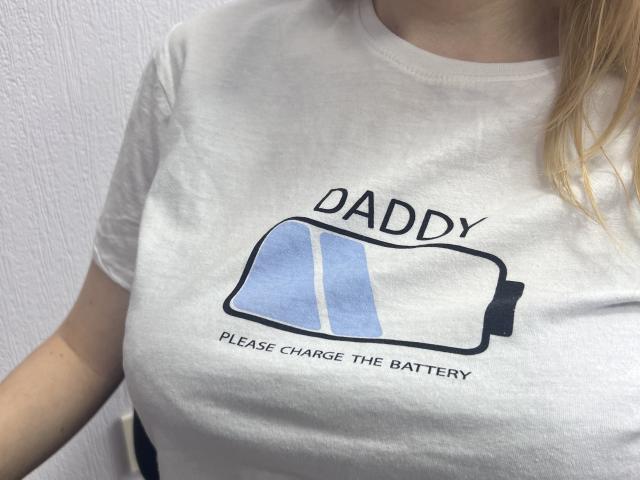 Laliina's white t-shirt with a battery graphic, text "DADDY" above and "PLEASE CHARGE THE BATTERY" below, covering her small, firm tits. Blonde hair on the right.