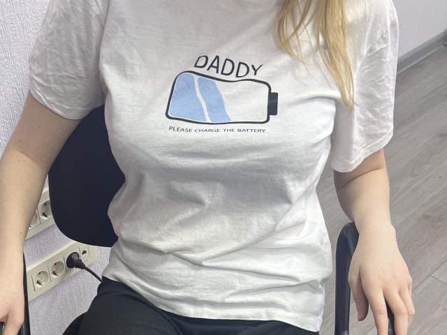 Laliina with pale skin, blonde hair, wearing a white t-shirt showing a battery with text, sitting on a black chair, hands on lap, in a room with white walls.