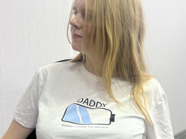 Laliina with long blonde hair, white t-shirt, and small tits, sitting and looking to the side with a subtle smile. Her face is slightly flushed.