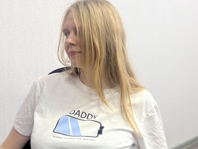 Laliina has long blonde hair covering her right eye, wearing a white T-shirt with a blue and black graphic, sitting against a white wall.