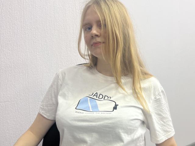 Laliina with long blonde hair, wearing a white t-shirt, small tits, sitting, light skin, subtle smile, white textured wall in background, casual, relaxed, slightly messy hair.
