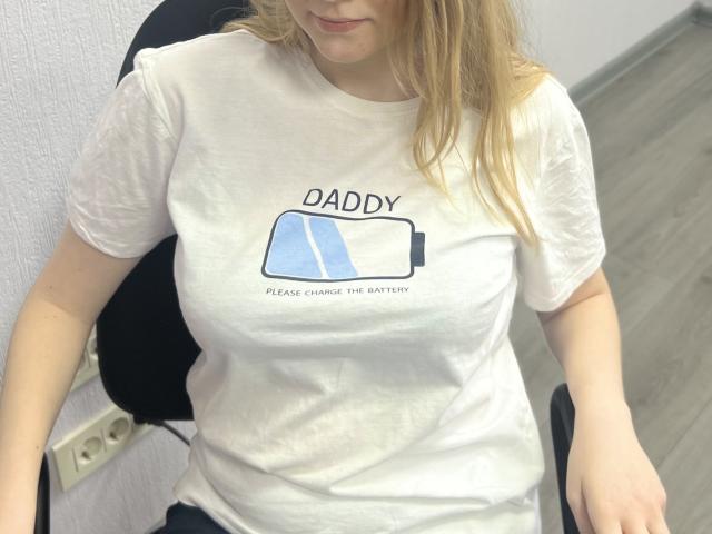 Laliina with blonde hair, white t-shirt, and large boobs, sitting in a black chair, wearing dark pants, in a room with gray wood flooring.
