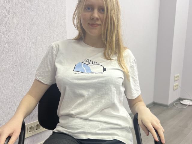 Laliina, blonde girl with fair skin, small tits, white t-shirt, sitting on black chair, smiling, gray walls, light wood floor, white wall plug visible, casual, relaxed.
