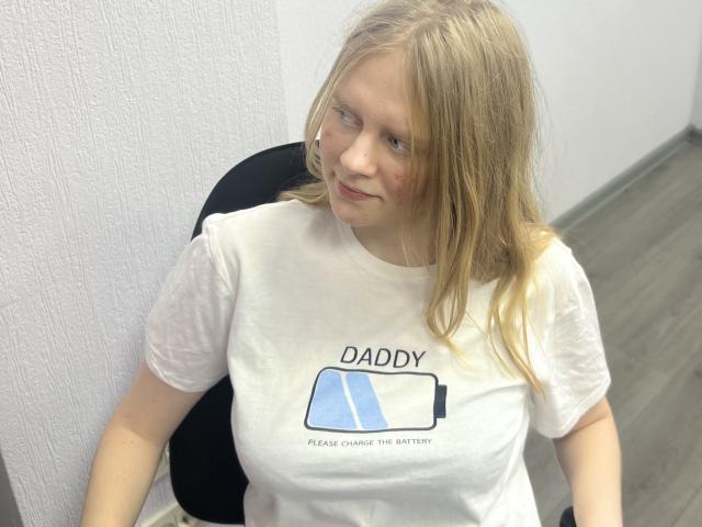 Laliina with blonde hair, small nose, white t-shirt, and "DADDY" shirt, sitting on black chair, wooden floor, and white textured wall, looking to the side with a slight smile.