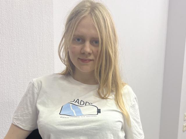 Laliina with long blonde hair, pale skin, small breasts, wearing a white T-shirt, standing against a white wall, looking slightly to the side with a subtle smile.