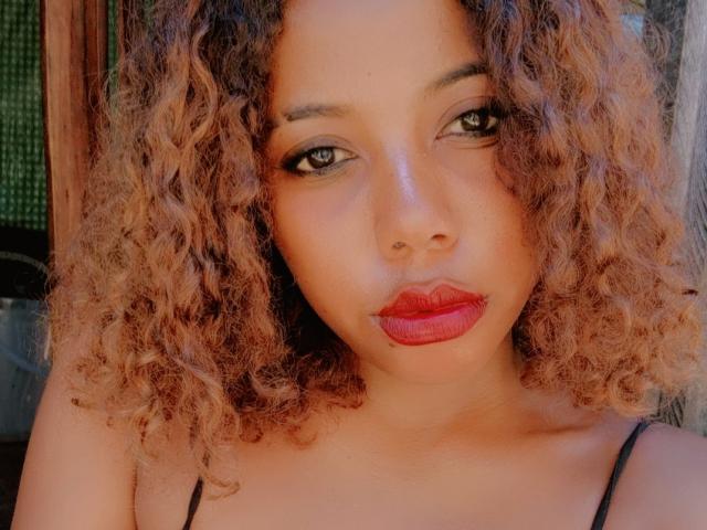 Daceselinn a les cheveux bouclés, d'un brun clair, du rouge à lèvres, et un eye-liner foncé, avec une expression sérieuse, regardant directement l'appareil photo. Ses yeux marrons ressortent sur sa peau claire..