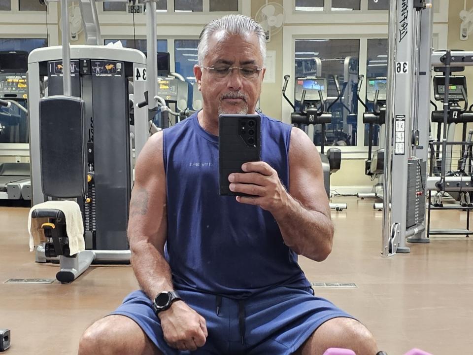 FlirtyOne4U, un hombre musculoso de mayor edad con cabello gris, gafas y una camiseta azul sin mangas, se toma un selfie en un gimnasio, sentado con las piernas separadas y un reloj negro..
